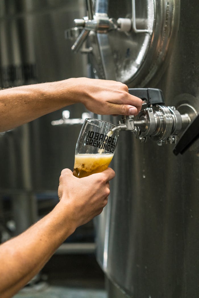 Close-up of beer being poured into a glass from a brewery tank by a person at a brewing facility.